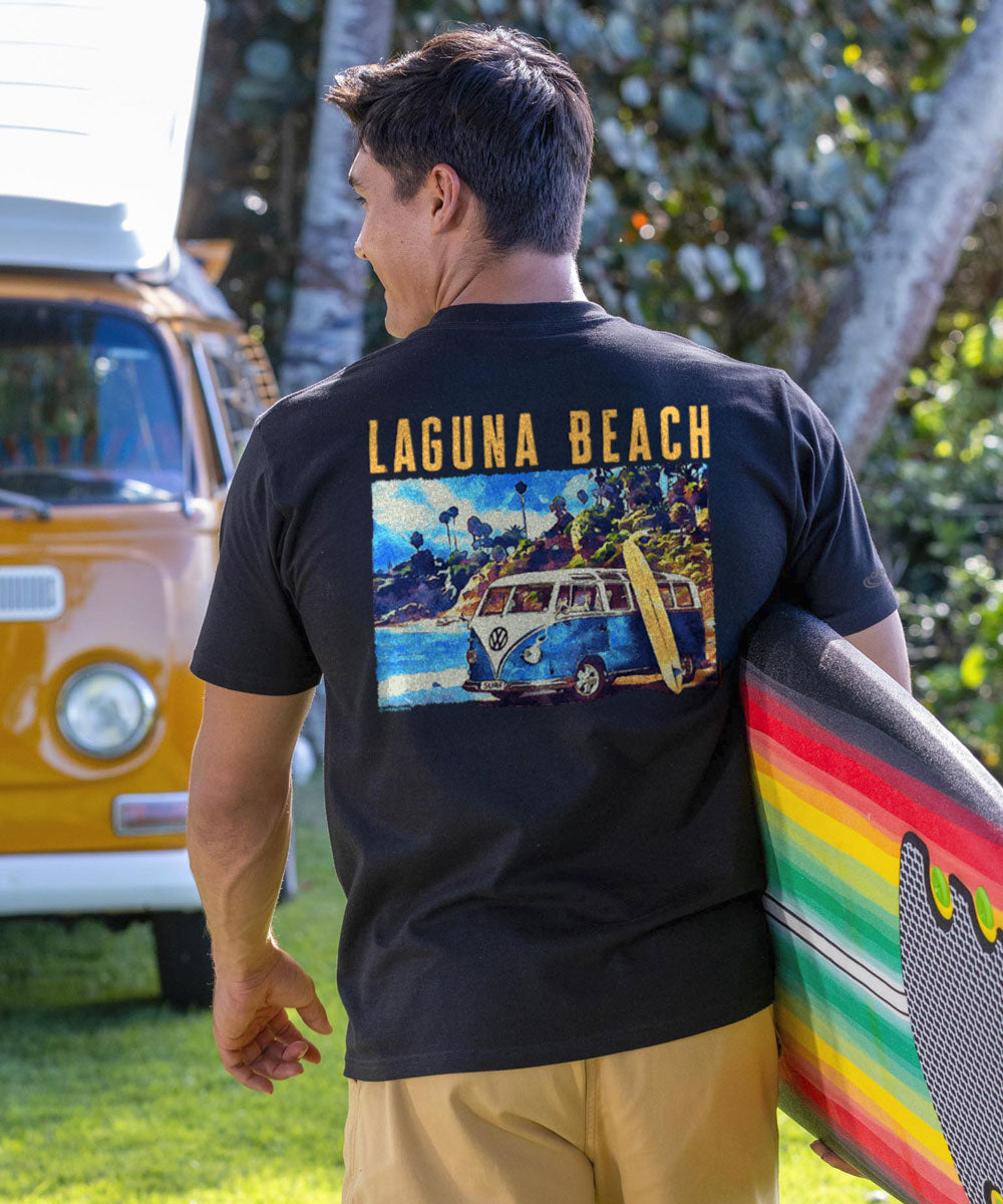 Man wearing a black t-shirt with 'Laguna Beach' design, holding a surfboard, with a vintage van and trees in the background. View 2