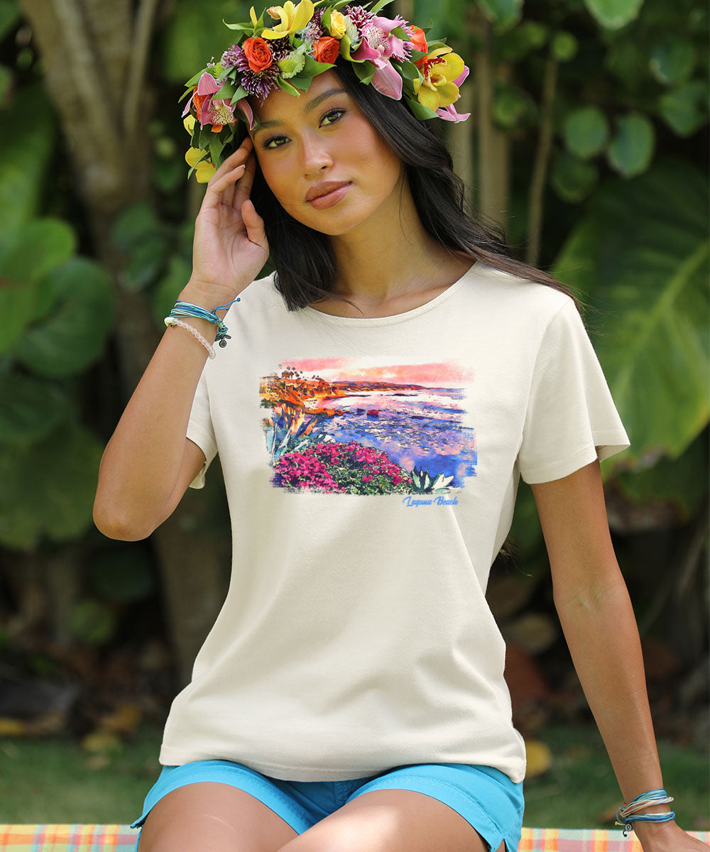 Woman wearing a white t-shirt with a colorful graphic design, sitting outdoors with greenery in the background. View 2