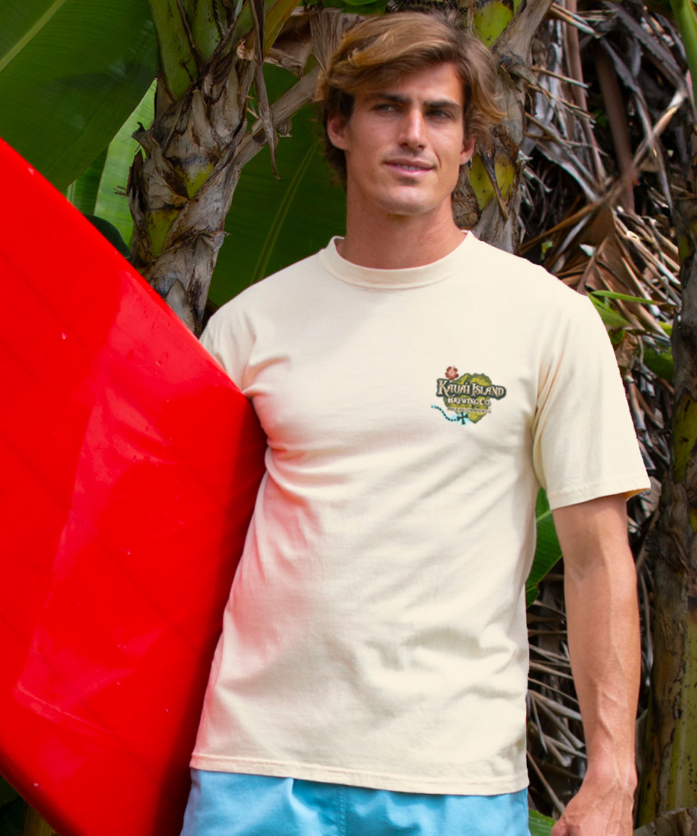 Man holding a red surfboard with a light beige t-shirt featuring a logo, standing in front of tropical plants. View 3