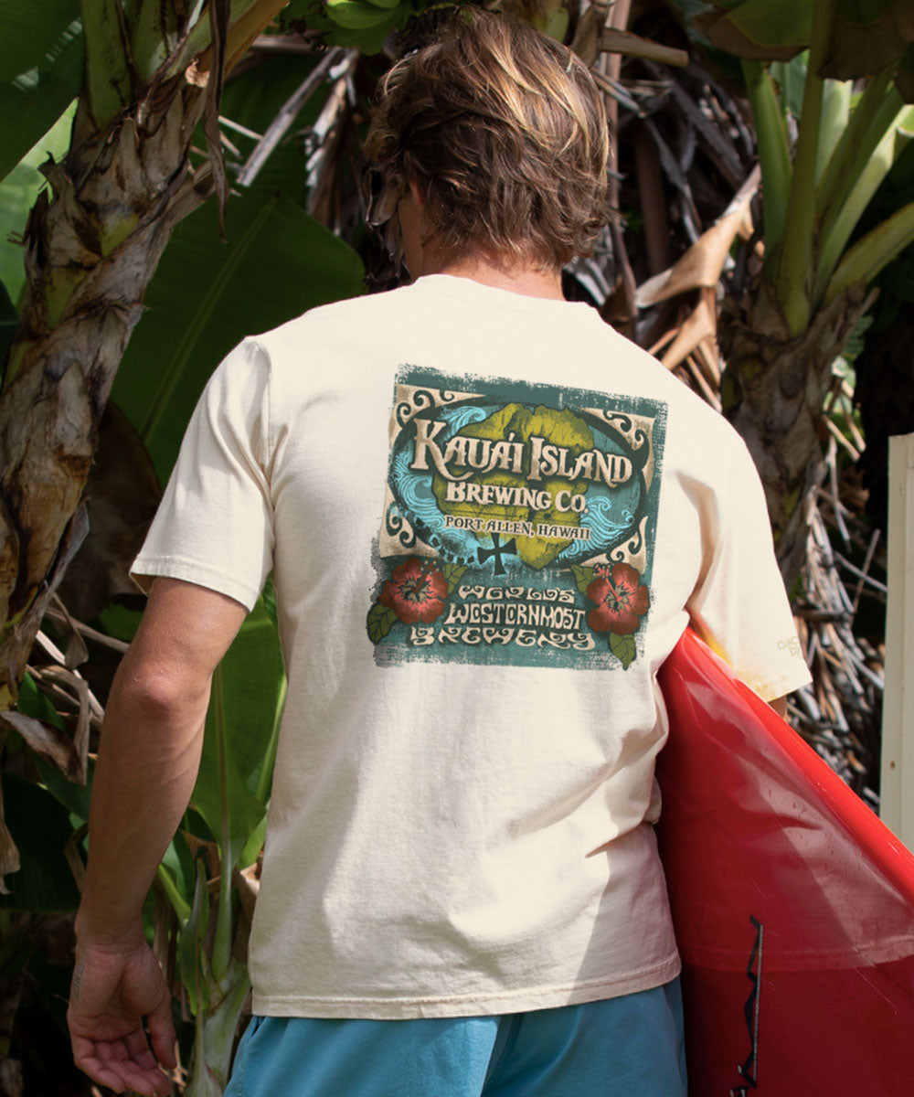 Man wearing a light beige t-shirt with Kauai Island Brewing Co. logo, holding a red surfboard. View 2