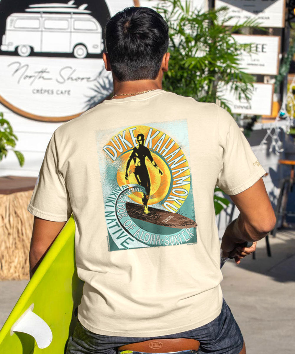 Person wearing a light beige t-shirt with a graphic design of a person on a surfboard, holding a surfboard. View 2