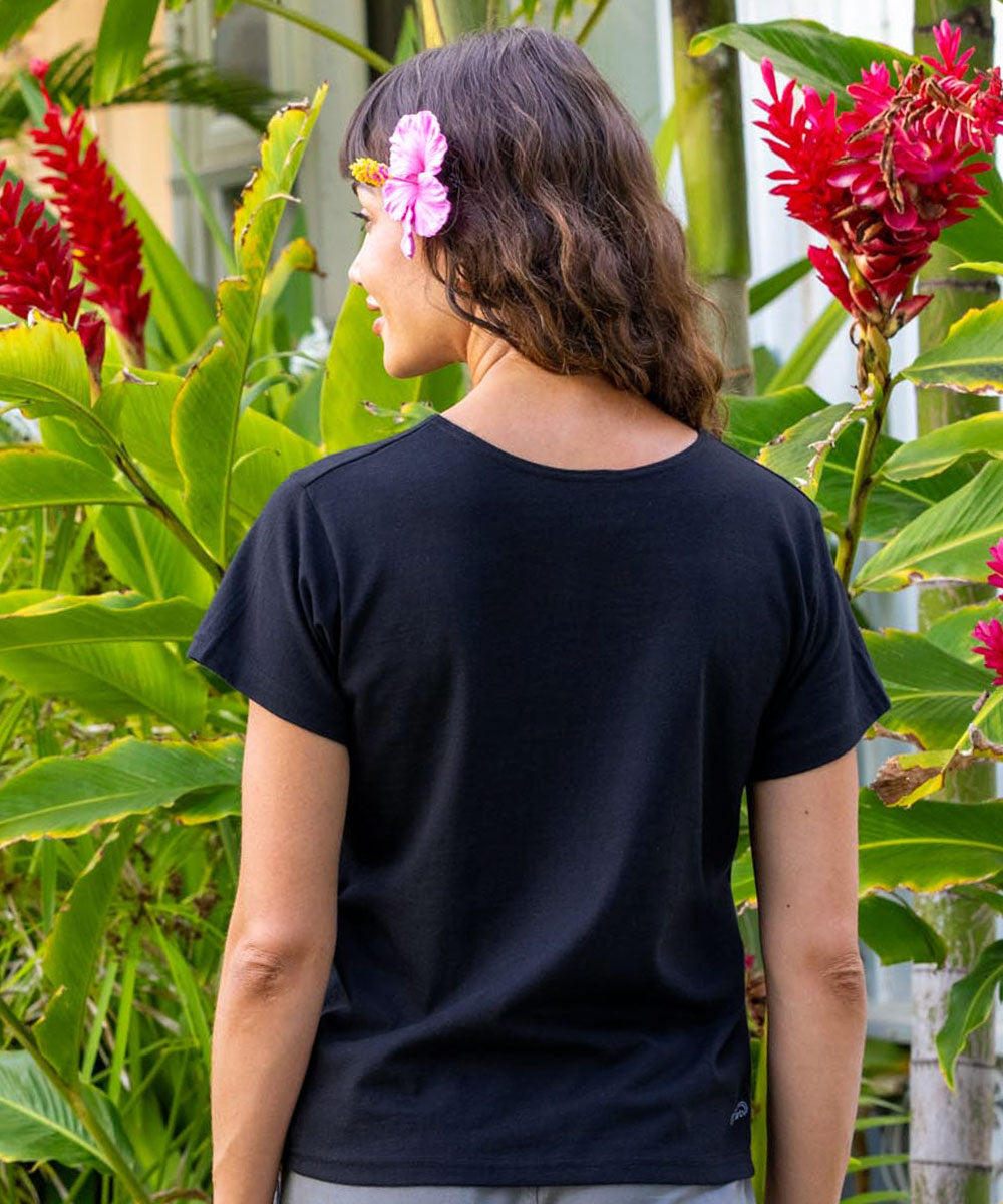 Person wearing a black t-shirt with flowers in hair standing among tropical plants View 3