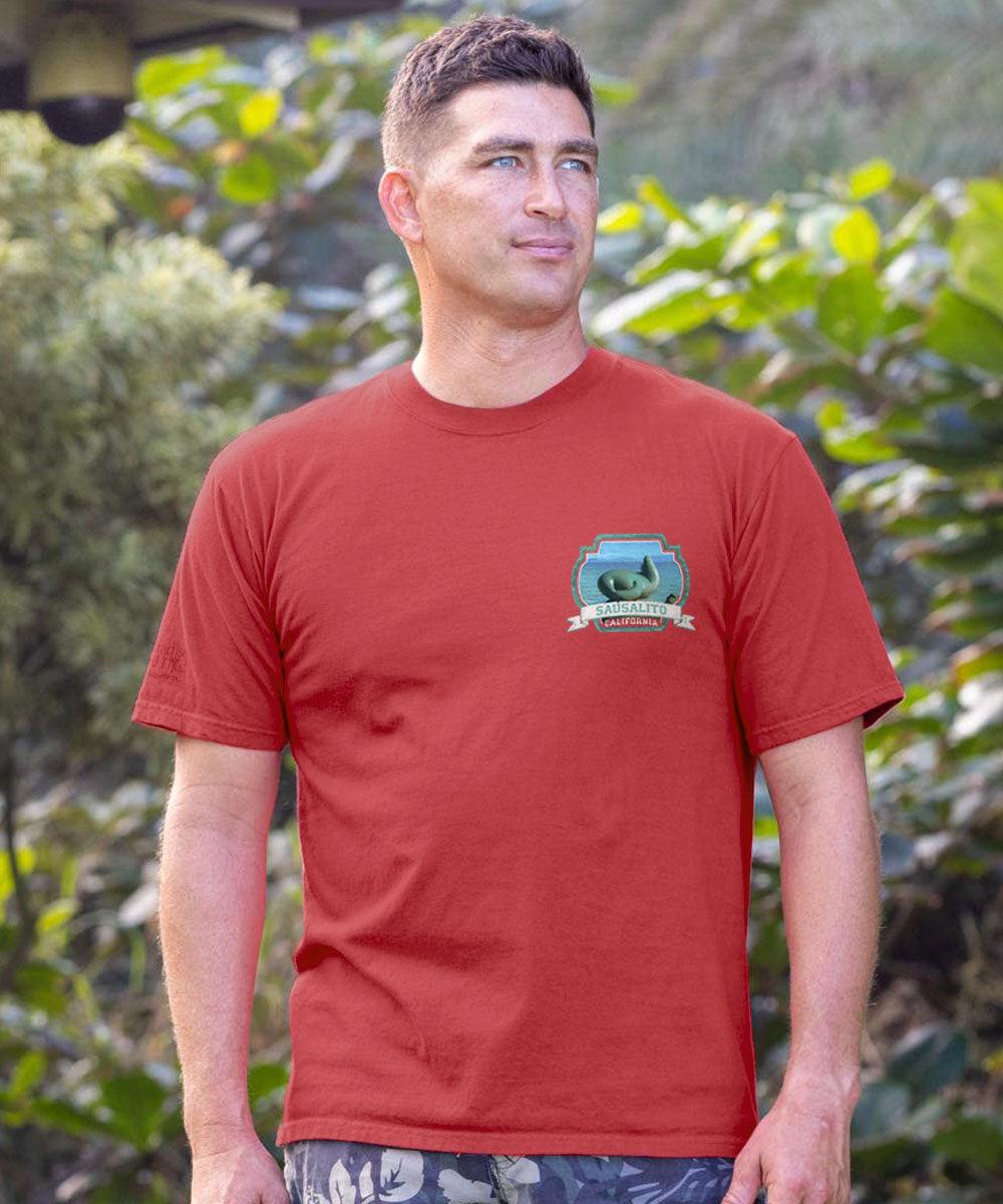 Man wearing a maroon t-shirt with a logo, standing in front of shrubs. View 3
