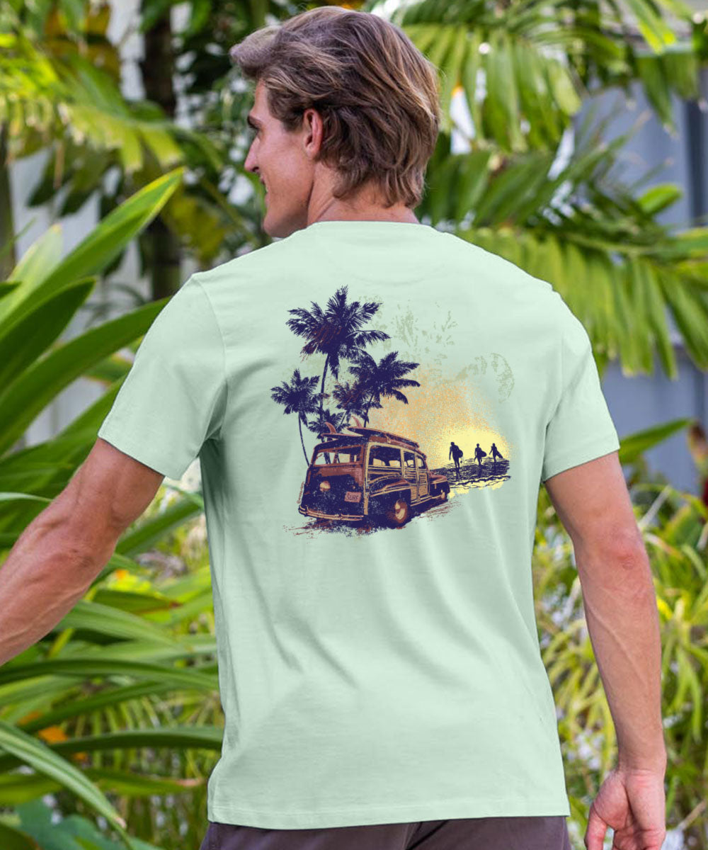 Man wearing a light green t-shirt with a palm tree and bus design, standing in front of green foliage. View 2