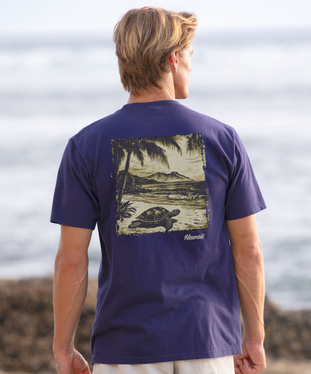Man wearing a blueish-purple t-shirt with a graphic of a turtle and palm trees, standing by the ocean. View 2