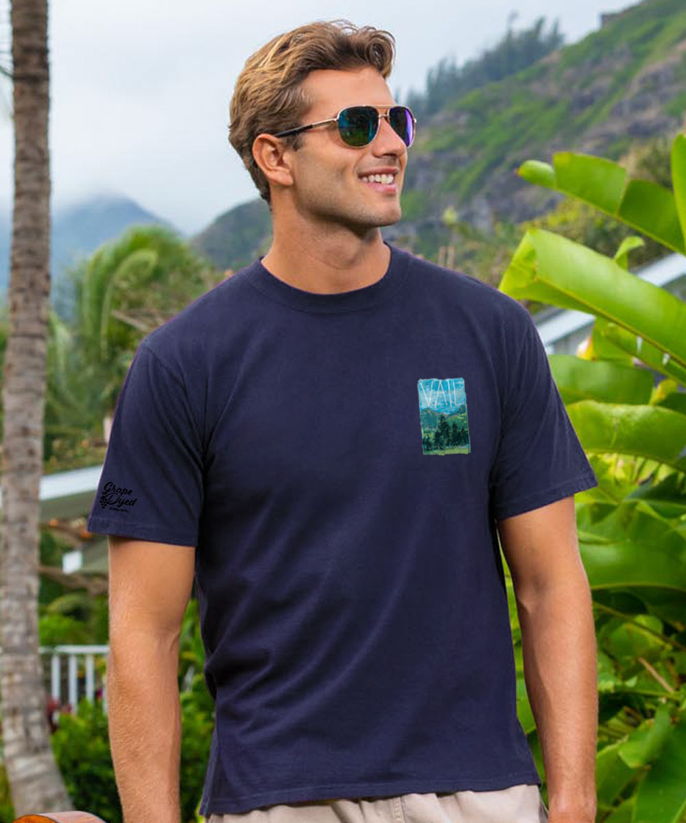 Man wearing a purple t-shirt with a pocket design, standing outdoors with greenery and mountains in the background. View 3