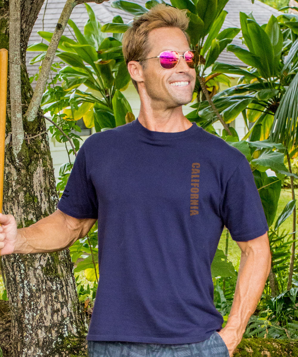 Man wearing a purple t-shirt with 'California' printed on it, standing outdoors among green plants. View 3