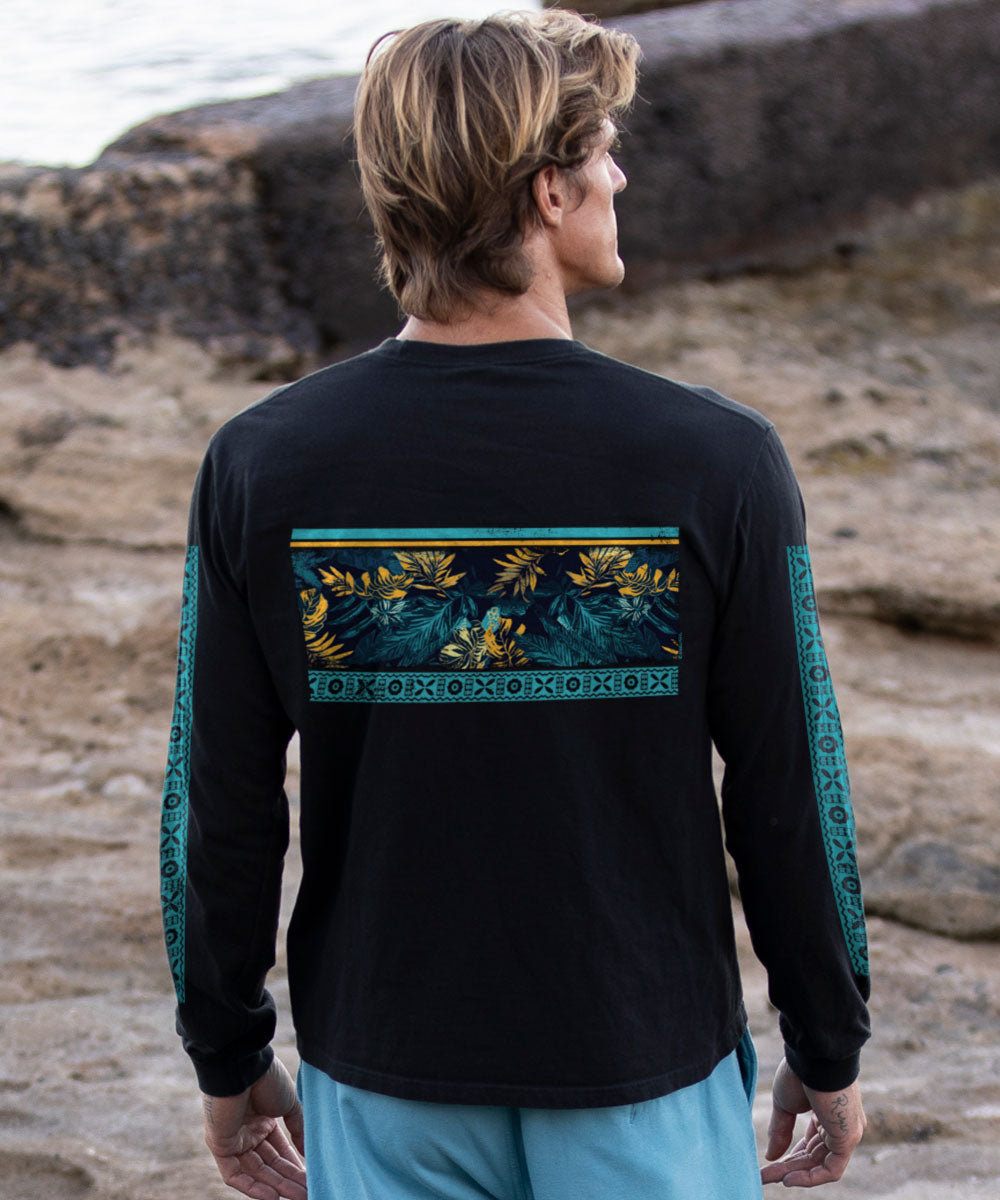 Man wearing a black long-sleeve shirt with floral leaf and geometric pattern design on the back, standing on a rocky beach. View 3