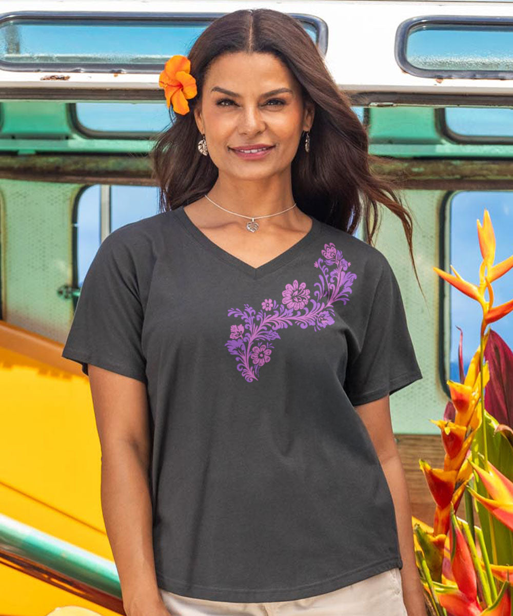 Woman wearing a black t-shirt with a purple floral design in front of a colorful bus and tropical plants. View 2