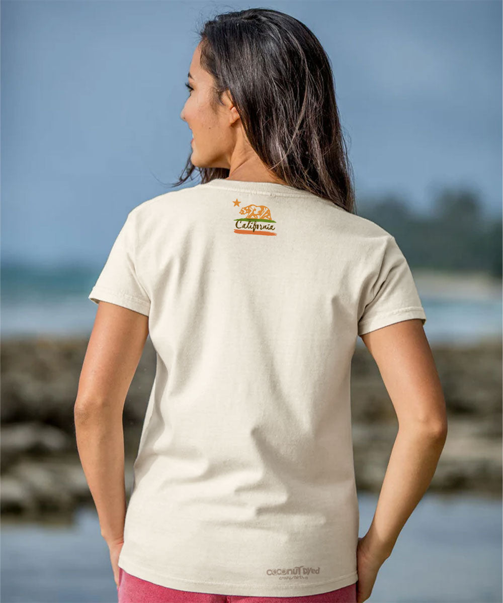 Woman wearing a beige t-shirt with a logo on the back, standing by a body of water. View 3