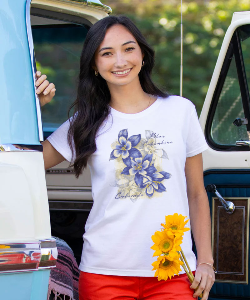Woman wearing a white t-shirt with a floral design, holding sunflowers, standing next to a vintage van. View 2