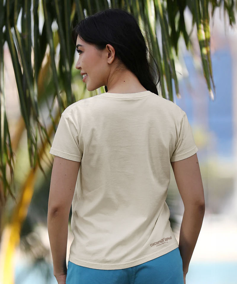 Woman wearing a beige t-shirt with a brand logo, standing outdoors with greenery in the background View 3