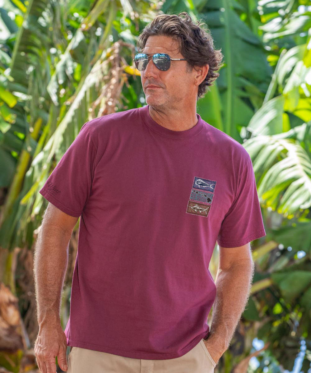 Man wearing a maroon t-shirt with a logo, standing in front of green foliage. View 3