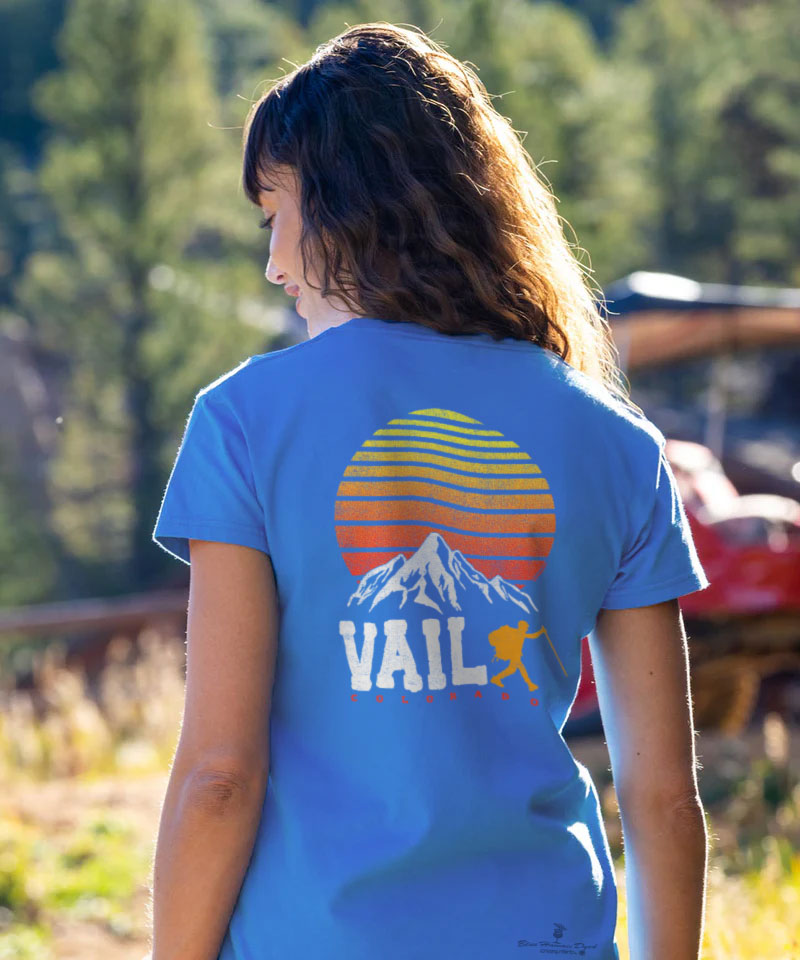 Person wearing a blue t-shirt with a mountain and 'Vail' design, standing outdoors. View 2