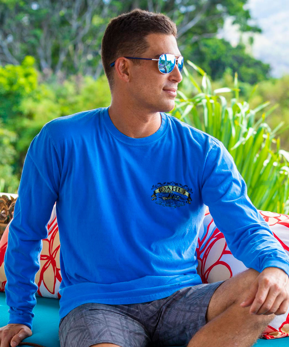 Man wearing a blue long-sleeve shirt with a logo, sitting outdoors with greenery in the background. View 3