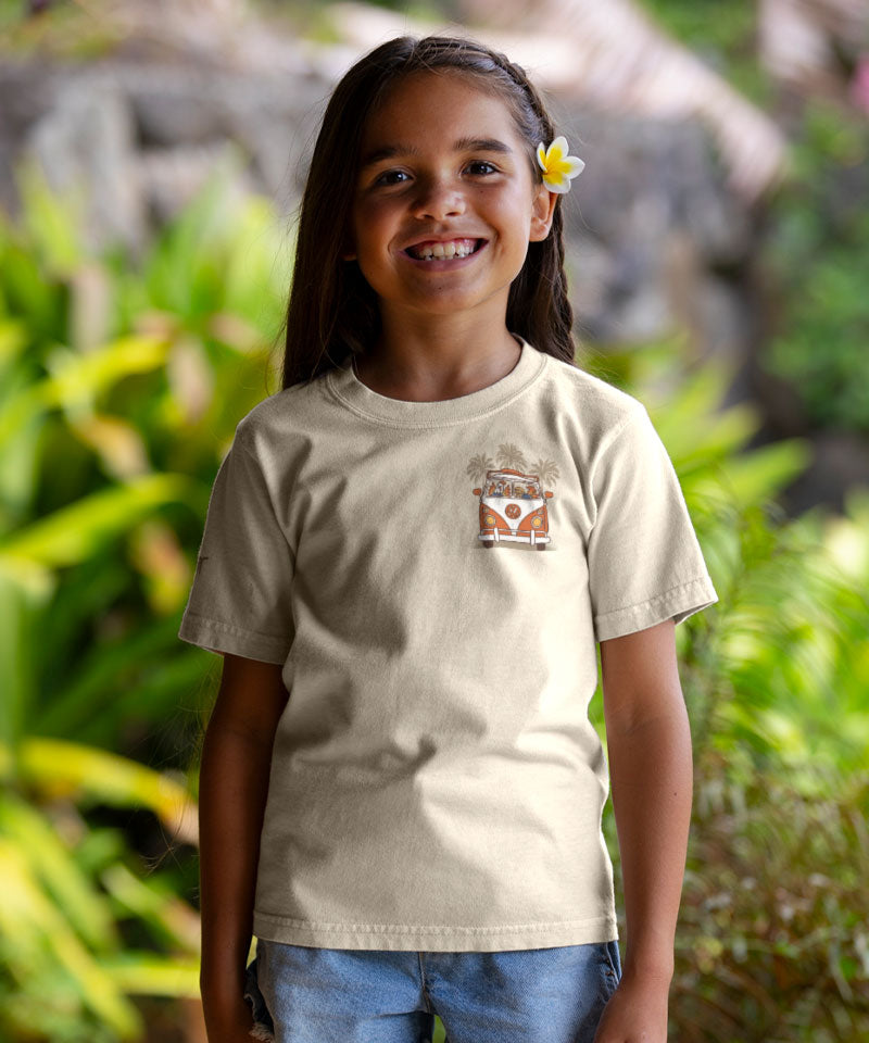 Young girl wearing a beige t-shirt with a logo, standing outdoors with greenery in the background View 3