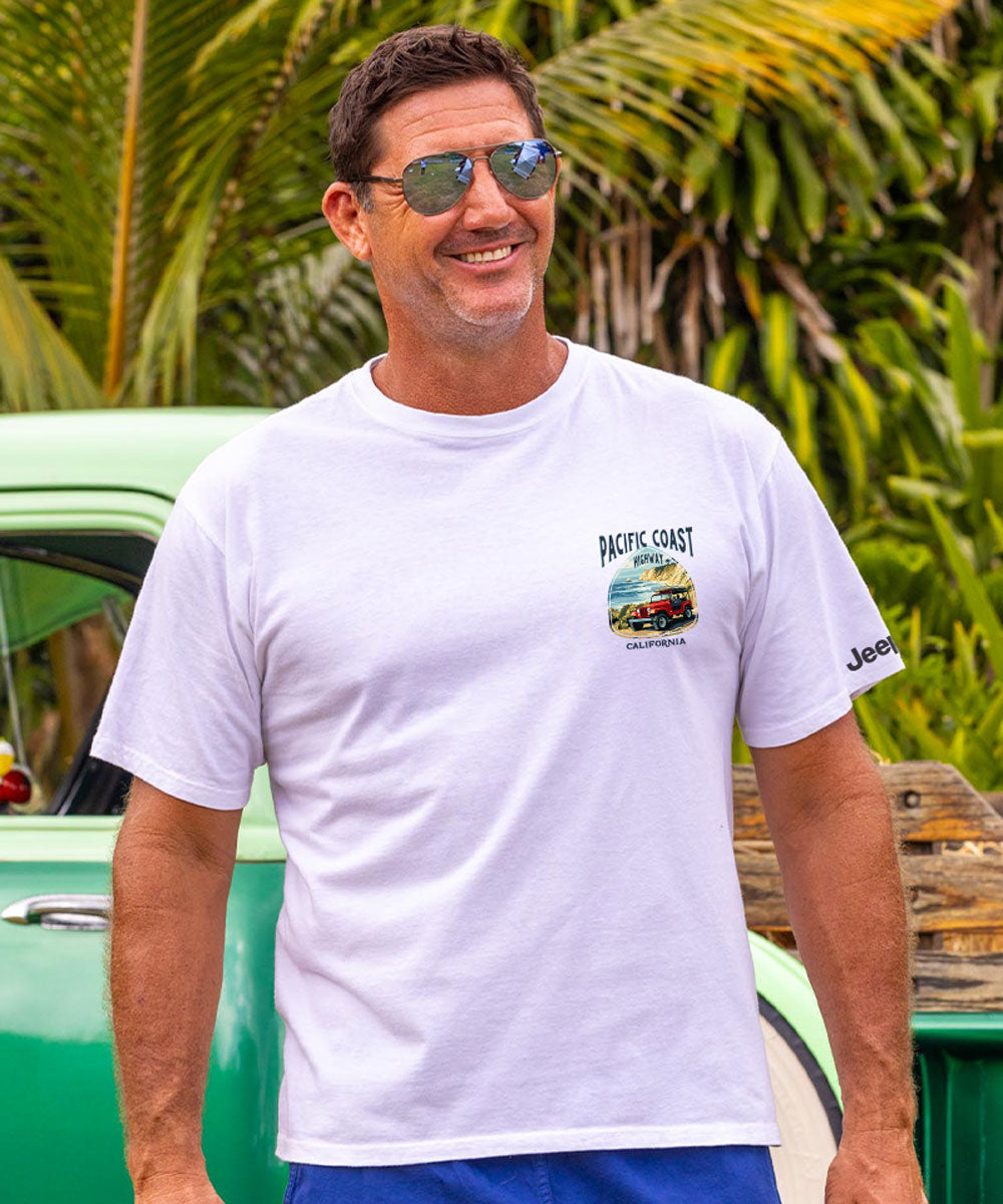 Man wearing a white t-shirt with a logo, standing in front of a green car with palm trees in the background. View 3