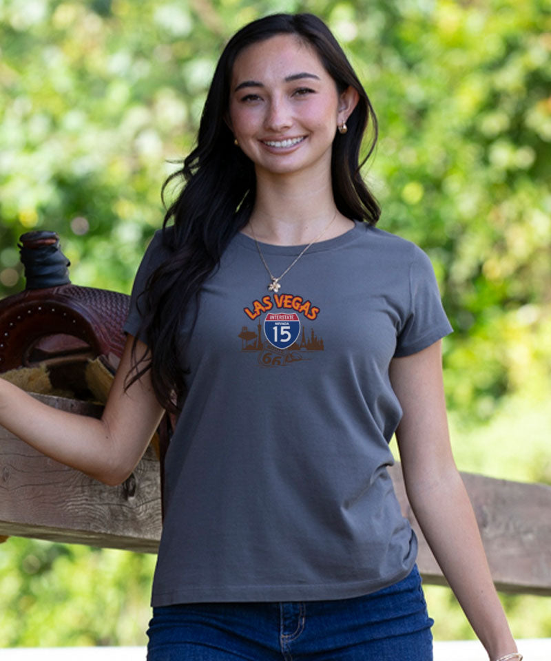 Woman wearing a gray t-shirt with Las Vegas design, standing outdoors with greenery in the background. View 3