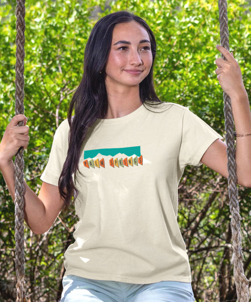 Woman wearing a t-shirt with a colorful graphic design, sitting on a swing with greenery in the background. View 3