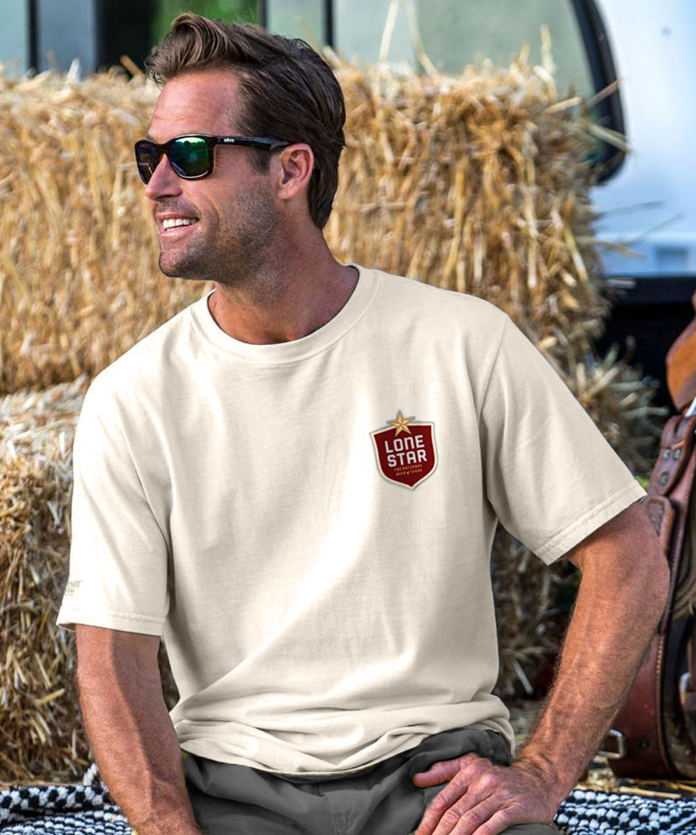 Man wearing a creamy white t-shirt with a logo sitting in front of hay bales. View 3