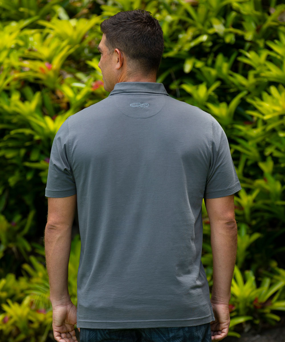 Man wearing a gray polo shirt with a patterned back design, standing in front of green foliage. View 3