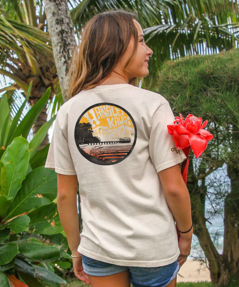 Woman wearing a t-shirt with a graphic design, standing outdoors with greenery and a beach in the background. View 2