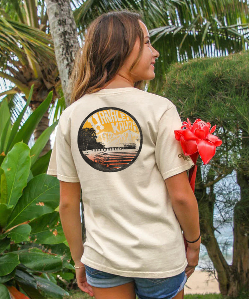Woman wearing a t-shirt with a graphic design, standing outdoors with greenery and a beach in the background. View 2