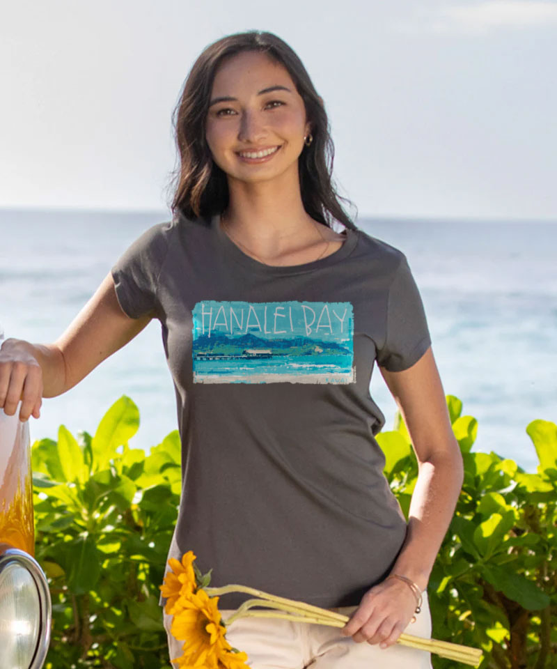 Woman wearing a gray t-shirt with a graphic design, standing outdoors with greenery and water in the background. View 2