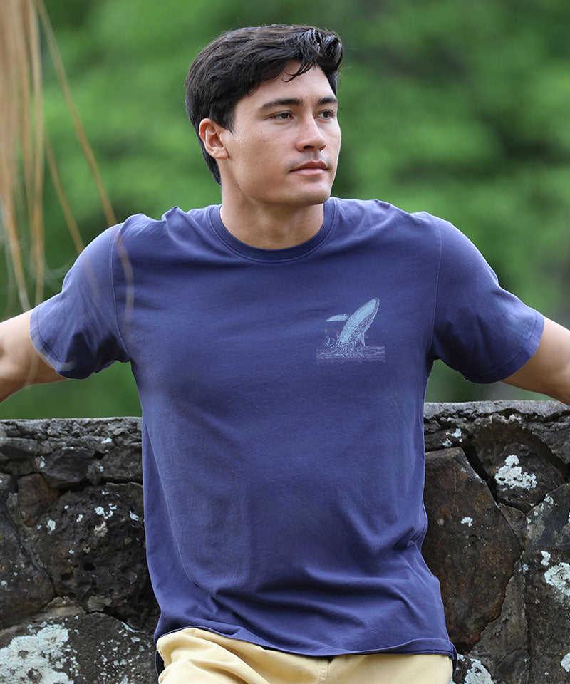 Man wearing an indigo t-shirt with a logo, standing outdoors against a stone wall View 3