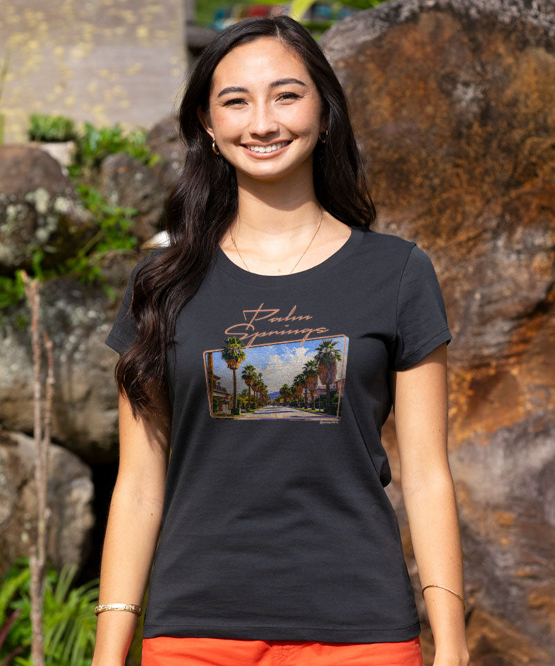 Woman wearing a black shirt with a 'palm springs' design on the front standing outside View 2