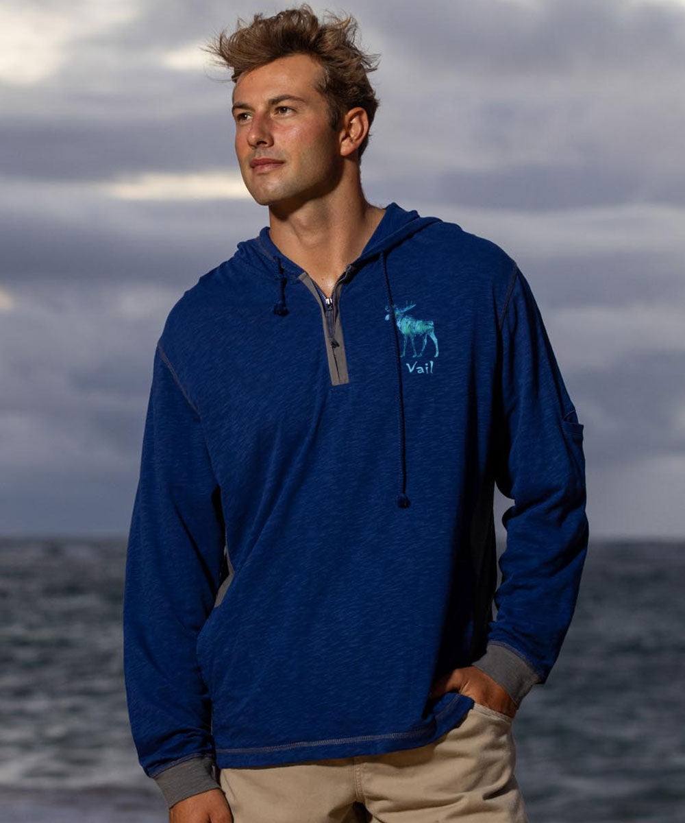 Man wearing a blue hoodie with a logo standing against a cloudy sky and ocean. View 3