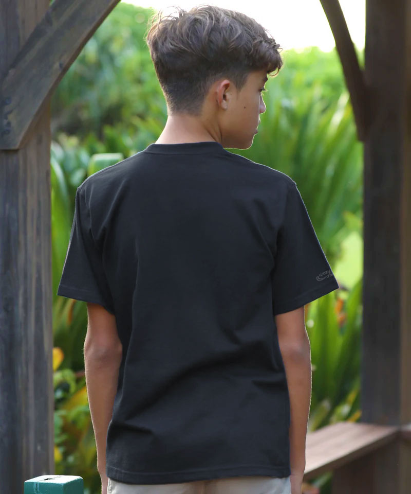 Person wearing a black t-shirt with a brand logo, standing outdoors with greenery in the background View 3