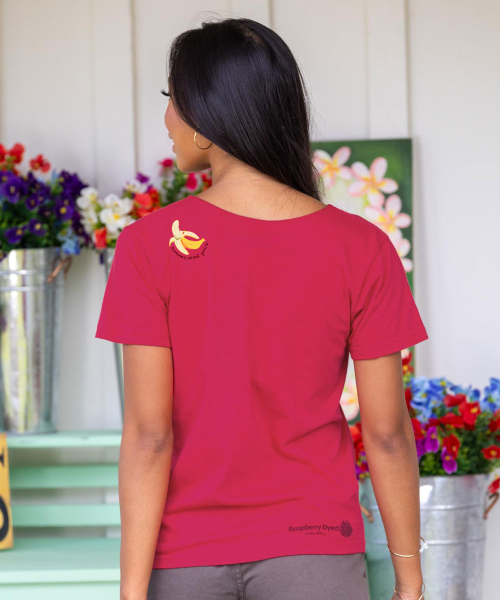 Person wearing a raspberry dyed t-shirt with a logo, standing in front of floral arrangements. View 3