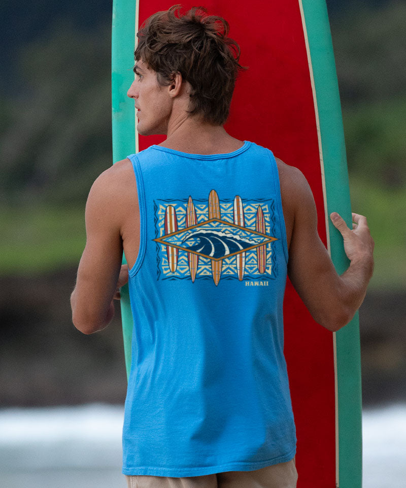 Man wearing a blue tank top with a colorful design on the back standing with a surfboard. View 2