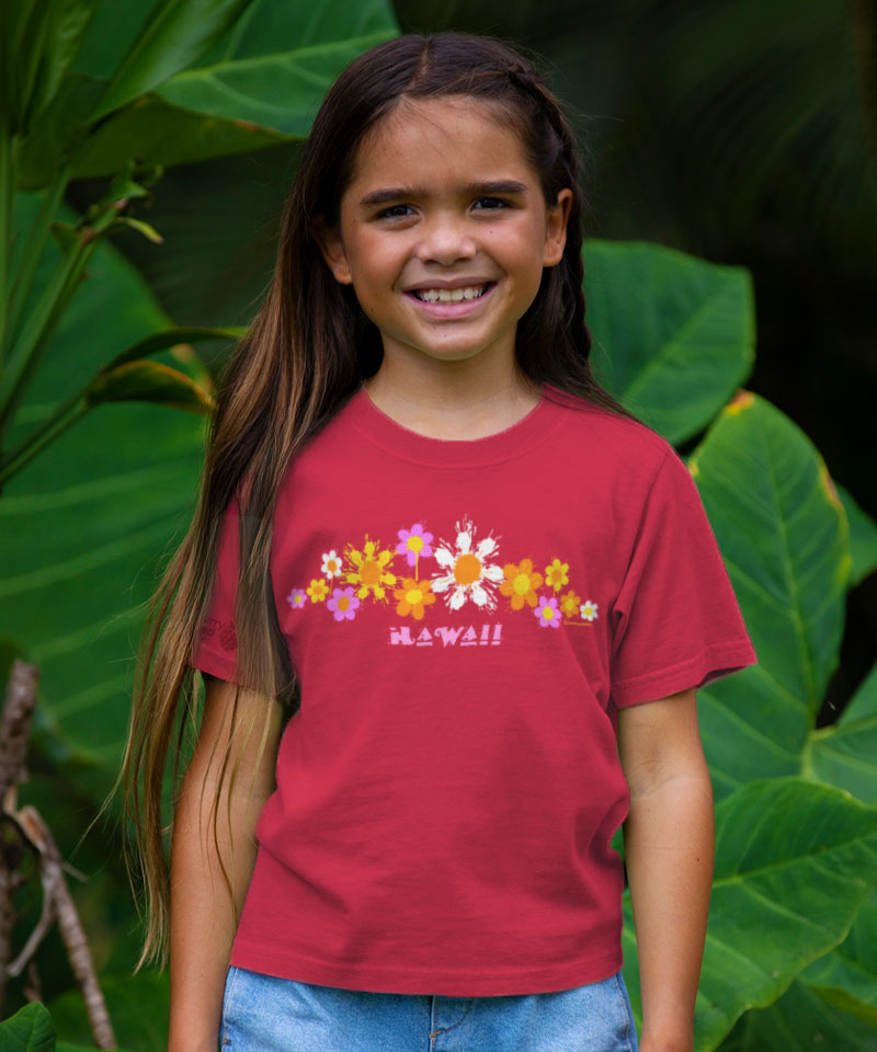 Young girl wearing a red t-shirt with floral designs and 'Hawaii' text, standing in front of green foliage. View 2