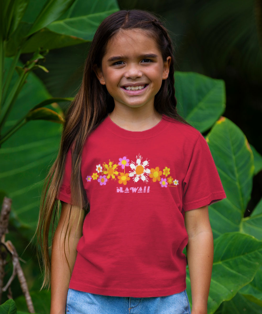 Young girl wearing a red t-shirt with floral designs and 'Hawaii' text, standing in front of green foliage. View 2