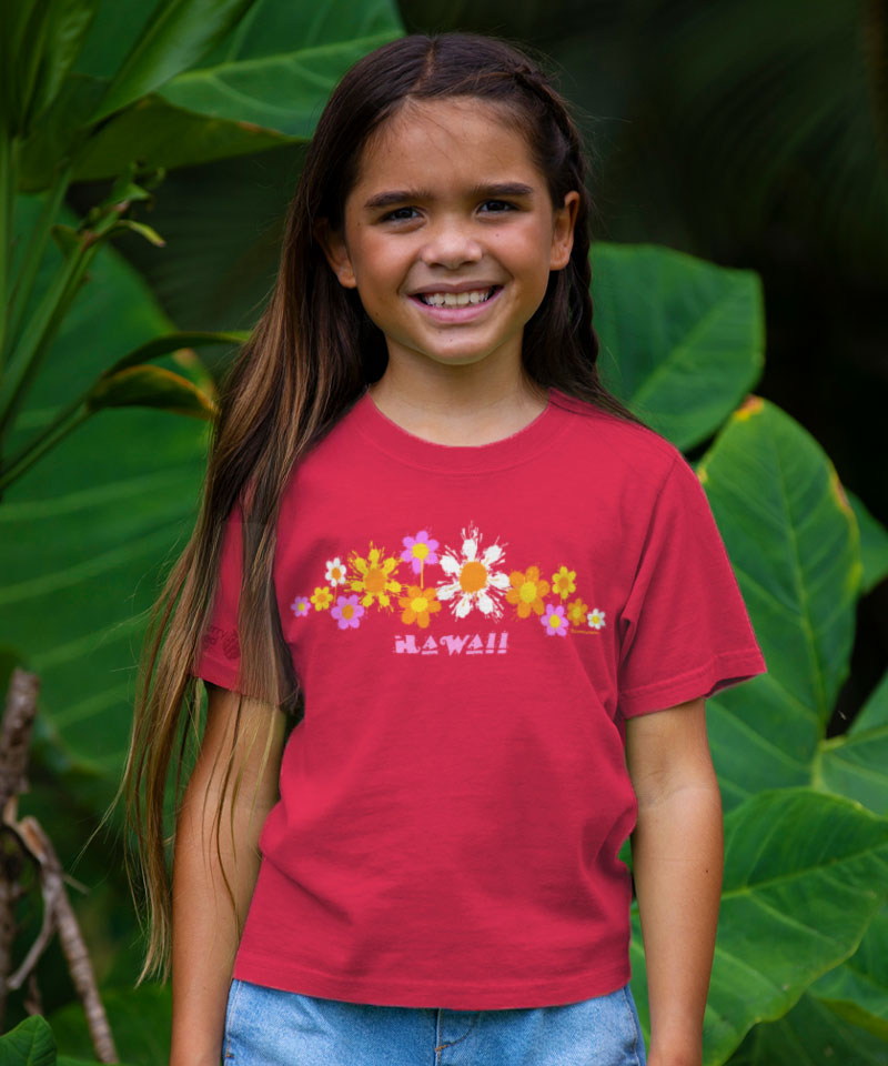Young girl wearing a red t-shirt with floral designs and 'Hawaii' text, standing in front of green foliage. View 2