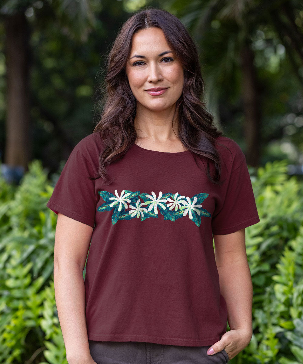 A woman with long dark brown hair stands outdoors in front of a blurred green foliage background, wearing a short-sleeve crewneck t-shirt in maroon with a horizontal graphic of white flowers and teal leaves across the chest. View 2