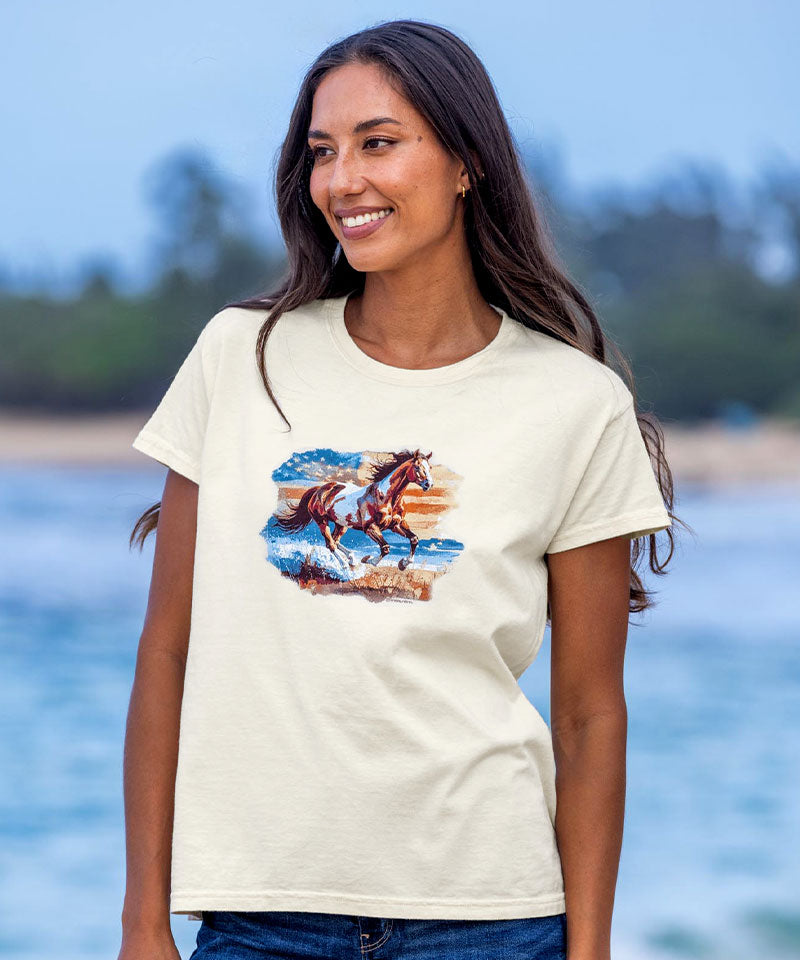 Woman wearing a t-shirt with a graphic of two horses running, standing by a body of water. View 2