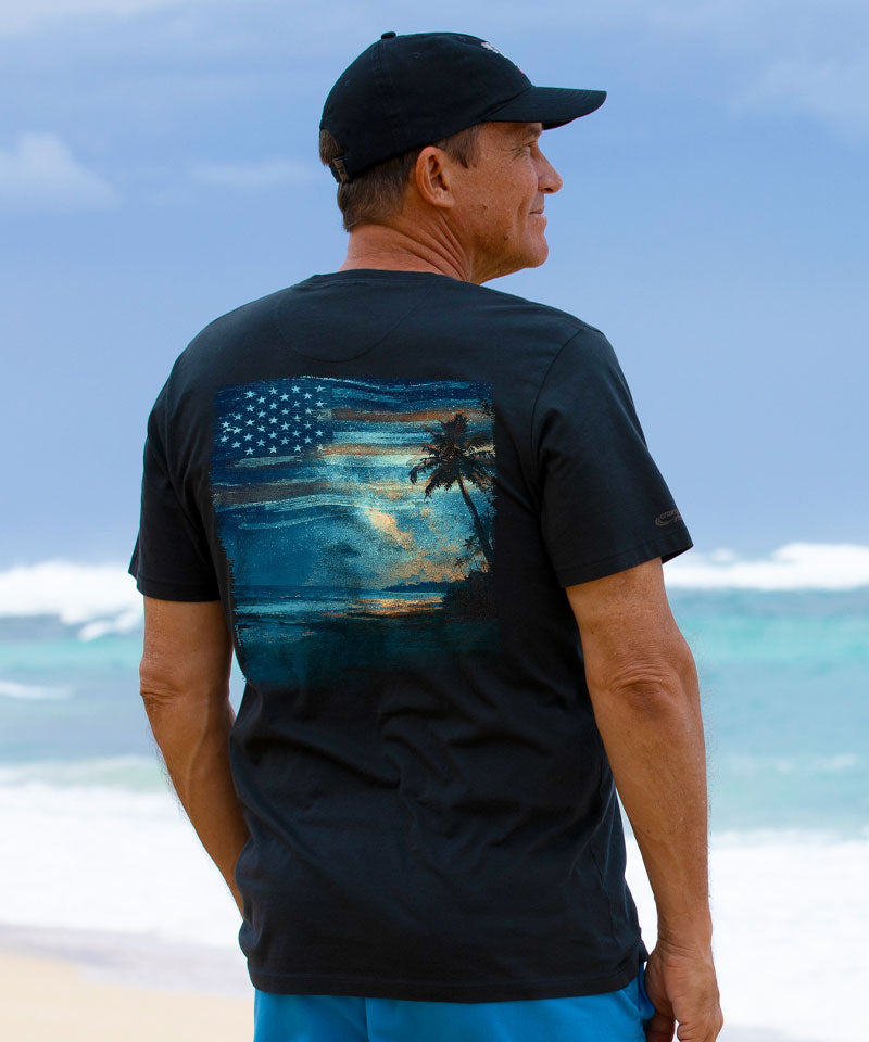 Man wearing a black t-shirt with a palm tree and American flag design on a beach. View 2