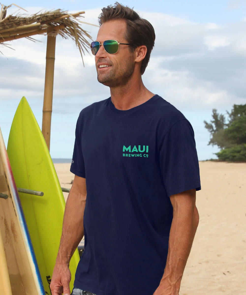 A man wearing a navy blue t-shirt with a green and white 'MAUI BREWING CO' logo on the chest, standing on a sandy beach with surfboards and a thatched umbrella in the background. View 3