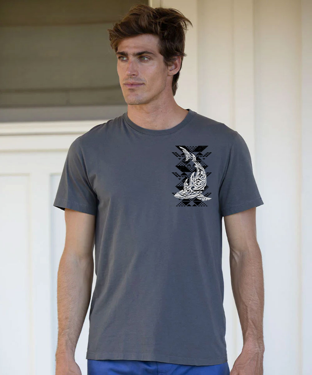 A man wearing a dark gray crewneck t-shirt with a black and white shark graphic on the left chest, standing against a white door background. View 3
