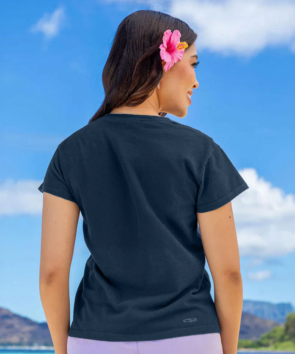 A woman shown from the back wearing a dark charcoal gray short-sleeve t-shirt, with a pink hibiscus flower in her hair, set against a bright blue sky and distant mountains. View 3