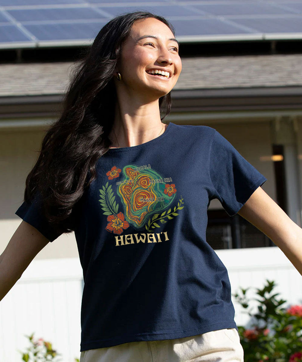 A woman wearing a navy blue graphic t-shirt with a colorful Hawaiian hula dancer design and the word 'HAWAII' printed in gold, standing outdoors in front of a house with solar panels. View 2