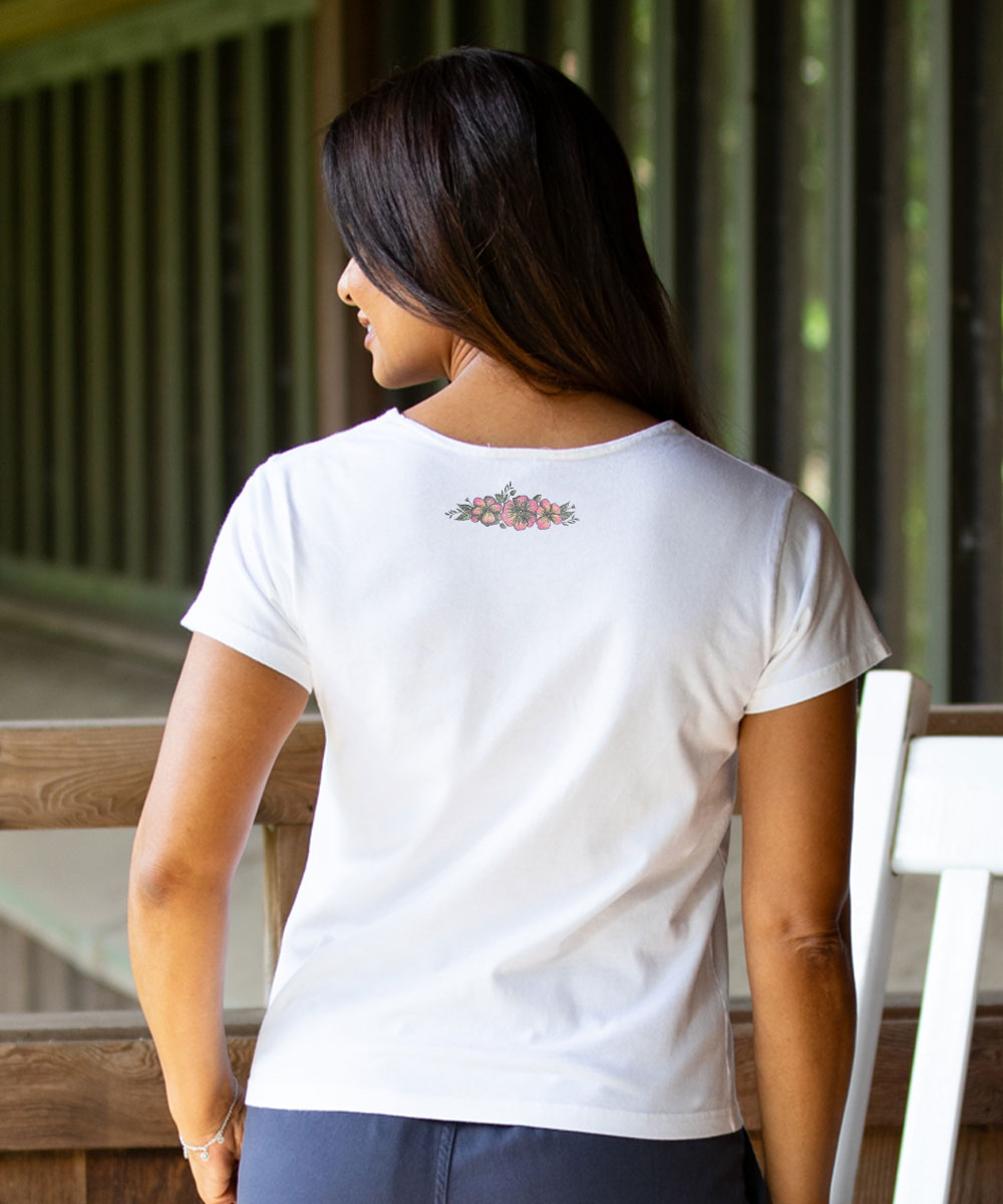 A woman viewed from behind wearing a white short-sleeve crewneck t-shirt with a small floral graphic printed at the center of the back, set against a rustic wooden porch background. View 3