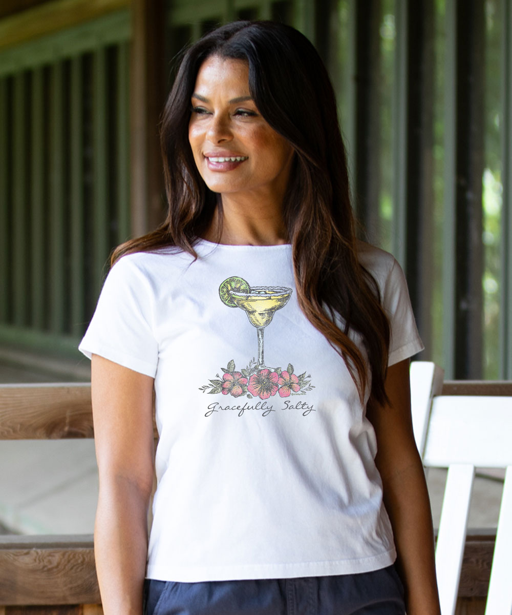 A woman wearing a white short-sleeve t-shirt with a graphic of a margarita glass, pink flowers, and the text 'Carefully, Sadly' printed on the front, set against a rustic wooden porch background. View 2