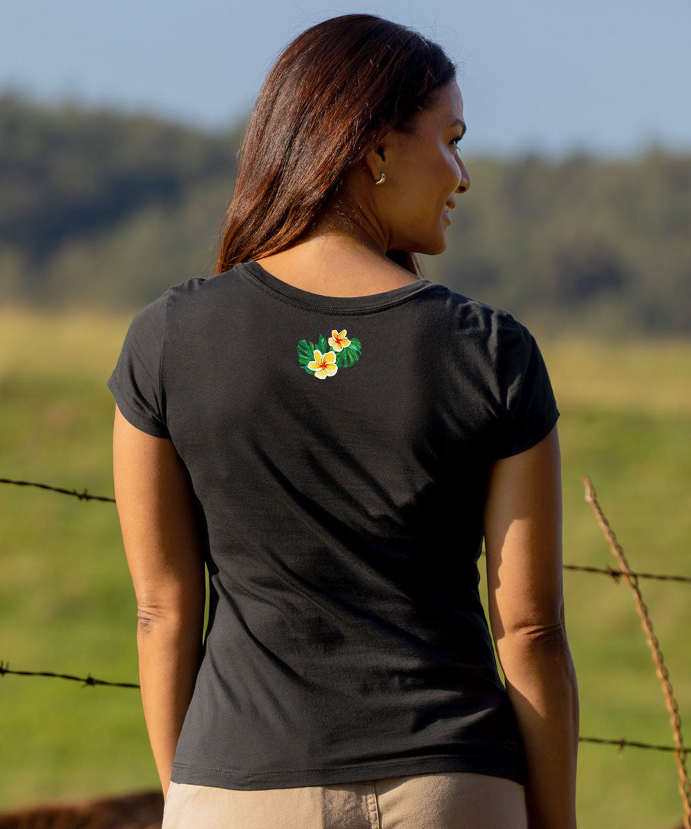 A woman wearing a black short-sleeve t-shirt with a small green and yellow floral graphic on the back of the neck, photographed outdoors in a grassy field. View 3