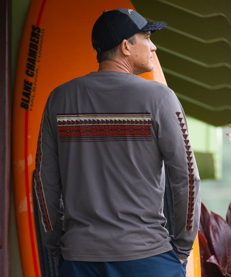 Man wearing a gray long-sleeve shirt with a patterned design, standing next to an orange surfboard. View 3