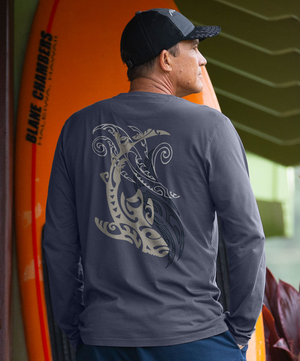 A man viewed from behind wearing a dark gray long-sleeve performance shirt with a large tribal Maori-inspired wave graphic on the back, paired with a black cap. He is standing next to an orange surfboard. View 2