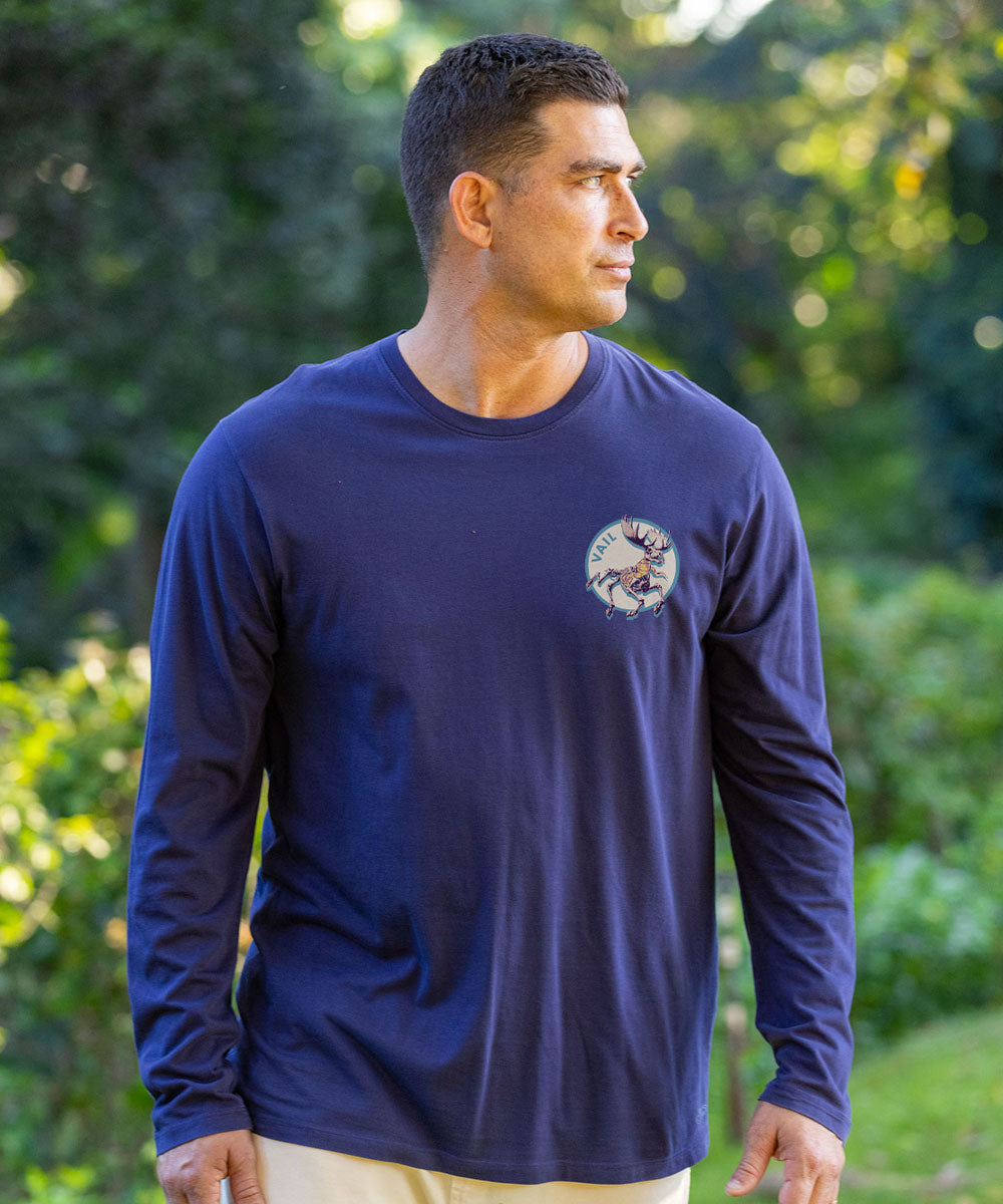 Man wearing a navy long-sleeve shirt with a logo, standing in front of a vintage car and tropical plants. View 3